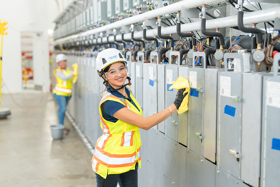 Female worker in safety gear smiles while cleaning electrical panels with a cloth. Another worker is visible in the background. Bright, professional setting.