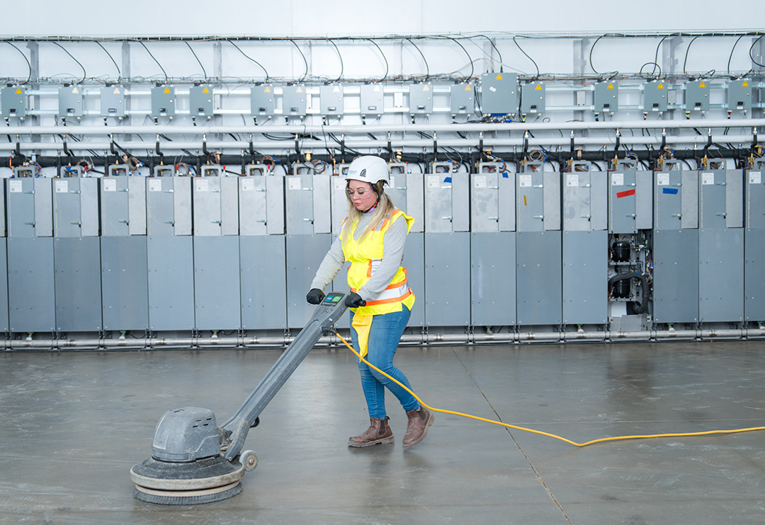 A woman in a hard hat and high-visibility vest operates an industrial floor buffer in a factory. Behind her are rows of electrical panels.