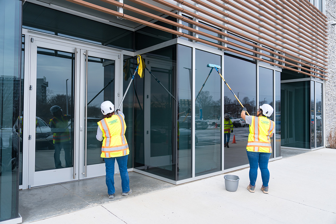 Two workers wearing hard hats and reflective vests clean large glass windows of a commercial building with mops. Scene conveys professionalism.