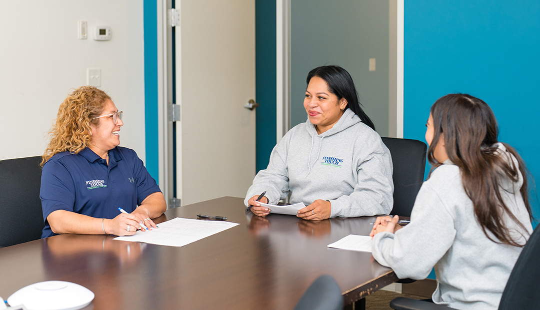 Three women sit at a conference table in a brightly lit room. They are smiling and holding papers, suggesting a collaborative, positive meeting.