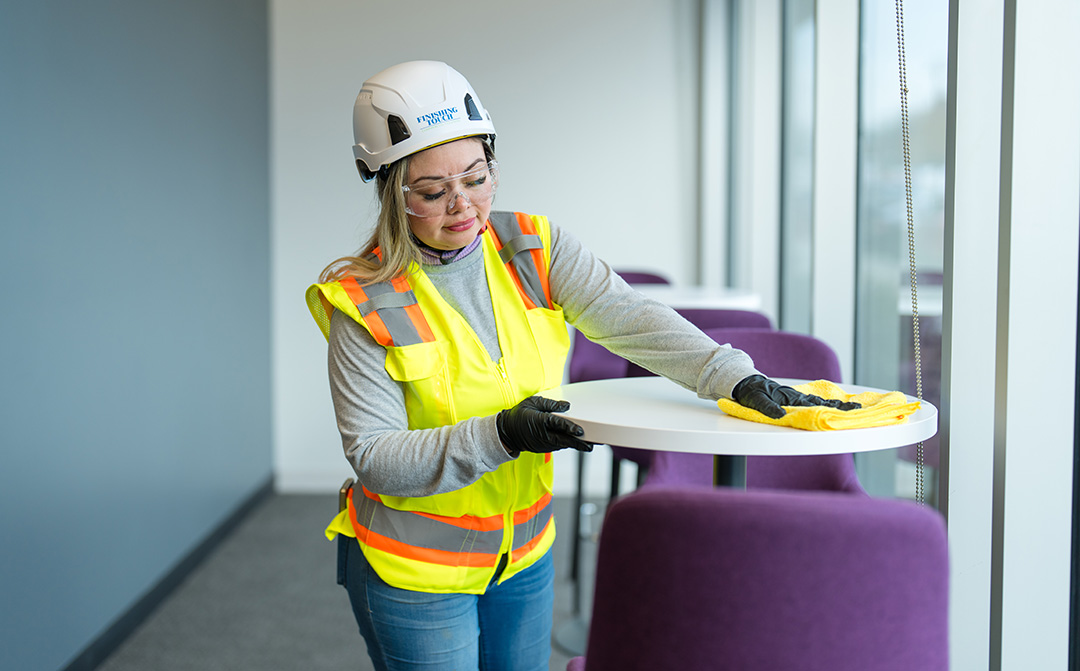 A woman in safety gear, including a helmet and high-visibility vest, is cleaning a white table with a yellow cloth in a modern, light-filled room.