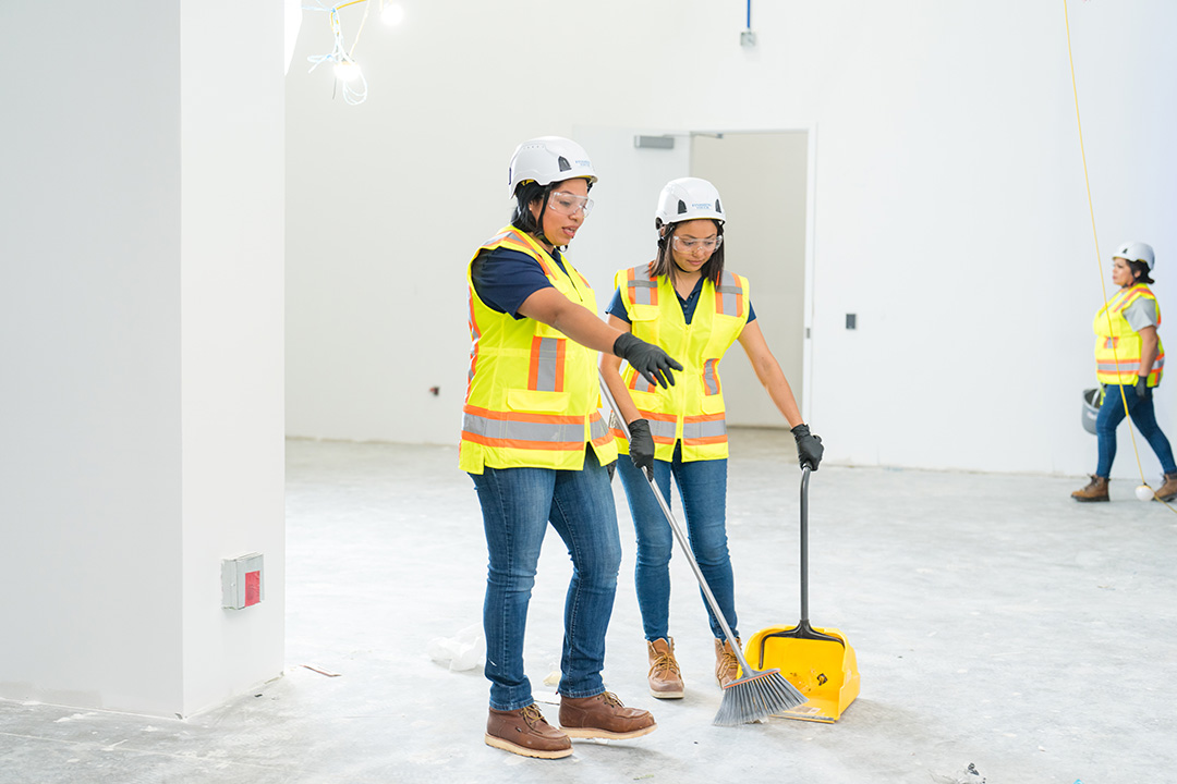 Two construction workers in safety gear and hard hats sweep a concrete floor in a bright, unfinished room. A third worker is in the background, creating a collaborative atmosphere.